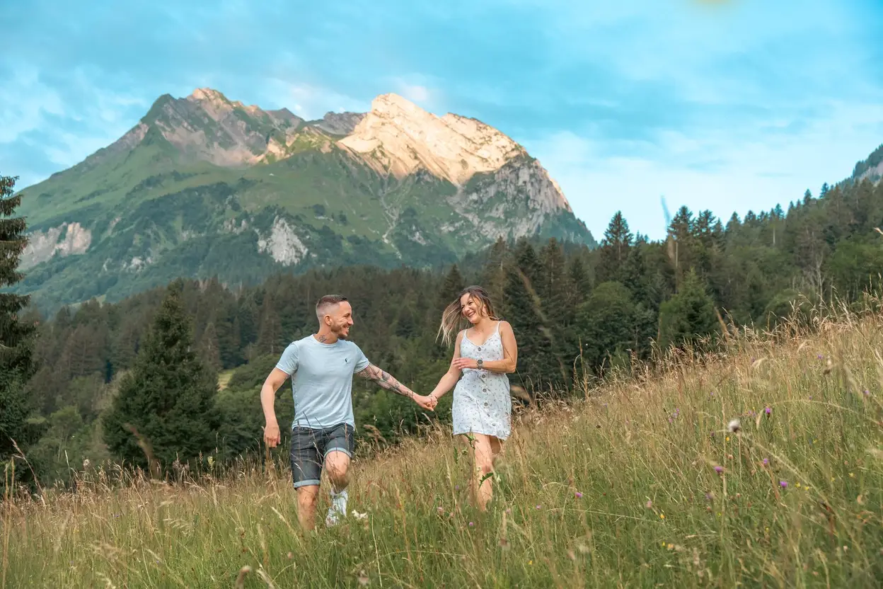 Verlobungsshooting in der Ostschweiz – Paar rennt lachend durch ein Feld vor Bergkulisse